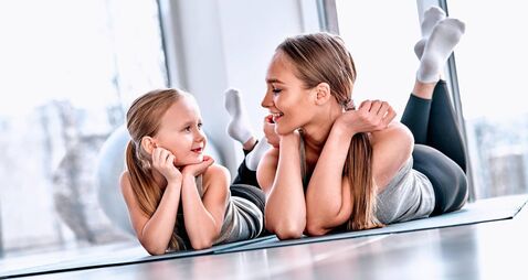 Beautiful young woman and charming little girl are looking at camera and smiling while lying on yoga mat in fitness hall