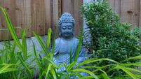 A close-up image of a gray stone Buddha statue sitting amidst a vibrant garden.
